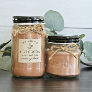 May include: Two glass jars with brown liquid inside, labeled "Old Fashioned Hot Cocoa served fresh daily warm up here". The jars are tied with twine and have black lids. The jars are sitting on a wooden surface with green leaves in the background.