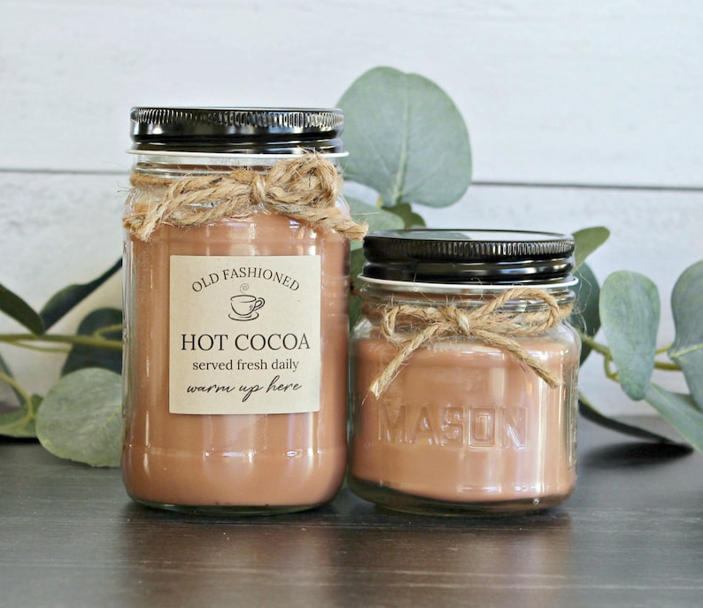 May include: Two glass jars with brown liquid inside, labeled "Old Fashioned Hot Cocoa served fresh daily warm up here". The jars are tied with twine and have black lids. The jars are sitting on a wooden surface with green leaves in the background.
