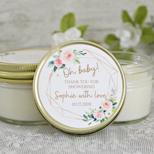 May include: A small glass jar candle with a gold lid and a white label with pink and green floral accents. The label reads "Oh baby! Thank you for showering Sophie with love 02.17.2019".