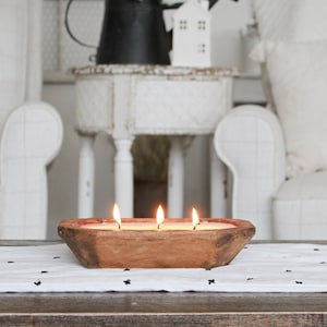 May include: A rectangular, hand-carved wooden dough bowl candle featuring three lit wicks. The natural brown wood has a weathered, rustic texture, and the white wax is filled to the brim of the vessel, creating a warm glow from the three flames.