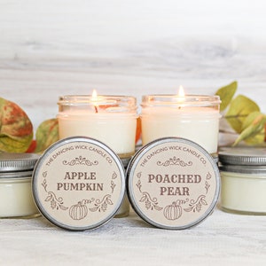 May include: Two lit candles in glass jars with metal lids, labeled "Apple Pumpkin" and "Poached Pear." Two additional candles with lids are visible. The Dancing Wick Candle Co. logo is on the labels. Autumn leaves are in the background.