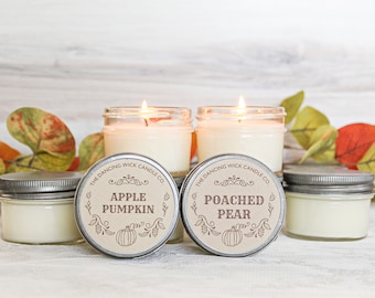 May include: Two lit candles in glass jars with metal lids, labeled "Apple Pumpkin" and "Poached Pear." Two additional candles with lids are visible. The Dancing Wick Candle Co. logo is on the labels. Autumn leaves are in the background.