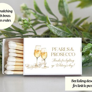 May include: White matchbox with a gold design featuring two glasses of champagne and pearls. The text on the box reads "Pearls & Prosecco" and "Thanks for lighting up Whitney's day!"
