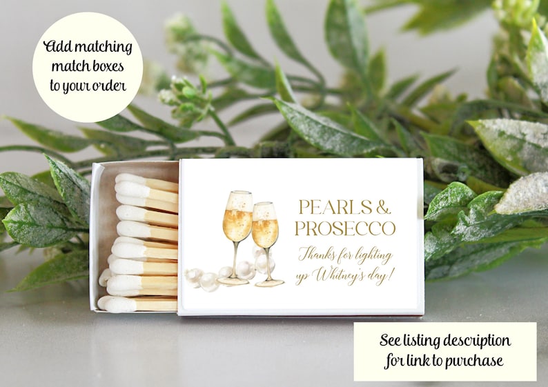 May include: White matchbox with a gold design featuring two glasses of champagne and pearls. The text on the box reads "Pearls & Prosecco" and "Thanks for lighting up Whitney's day!"