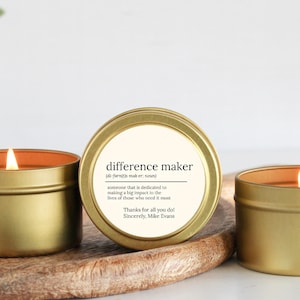 May include: Gold candle tin with a white label that reads "difference maker (di-farn(t)s mak-er, noun) someone that is dedicated to making a big impact in the lives of those who need it most Thanks for all you do! Sincerely, Mike Evans"