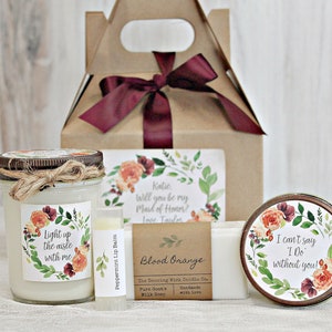 May include: A bridesmaid gift box with a floral theme. The box contains a candle, a bar of soap, a lip balm, and a small tin with a floral design. The candle label says "Light up the aisle with me". The soap label says "Blood Orange". The tin label says "I can't say I Do without you!"