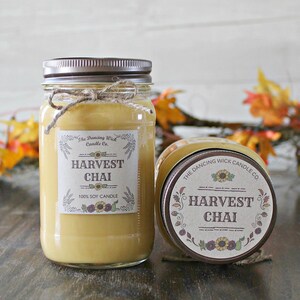 K&ouml;nnte beinhalten: Zwei Glasbeh&auml;lter mit braunen Deckeln und Etiketten. Die Etiketten sind cremefarben mit braunem Text und einem Sonnenblumendesign. Der Text auf den Etiketten lautet "The Dancing Wick Candle Co." und "Harvest Chai 100% Soy Candle".