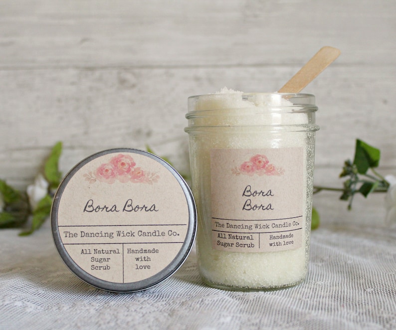 May include: A glass jar filled with white sugar scrub with a wooden spoon inside. The jar has a brown label with the text "Bora Bora The Dancing Wick Candle Co. All Natural Sugar Scrub Handmade with love" and a floral design.