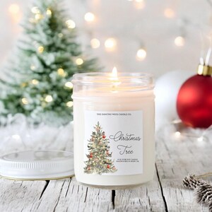 K&ouml;nnte beinhalten: Eine wei&szlig;e Kerze in einem durchsichtigen Glasgef&auml;&szlig; mit einem Etikett, auf dem "The Dancing Wick Candle Co. Christmas Tree 8 oz Pure Soy Candle Handpoured in Boise, ID" steht. Das Etikett zeigt eine Aquarell-Illustration eines Weihnachtsbaums mit Ornamenten und Lichtern.