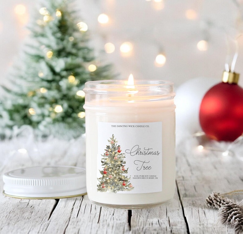K&ouml;nnte beinhalten: Eine wei&szlig;e Kerze in einem durchsichtigen Glasgef&auml;&szlig; mit einem Etikett, auf dem "The Dancing Wick Candle Co. Christmas Tree 8 oz Pure Soy Candle Handpoured in Boise, ID" steht. Das Etikett zeigt eine Aquarell-Illustration eines Weihnachtsbaums mit Ornamenten und Lichtern.