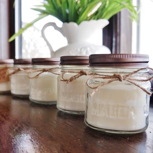 May include: Five glass jars with copper lids and twine tied around the top. The jars are filled with white wax and are arranged in a row on a wooden surface.
