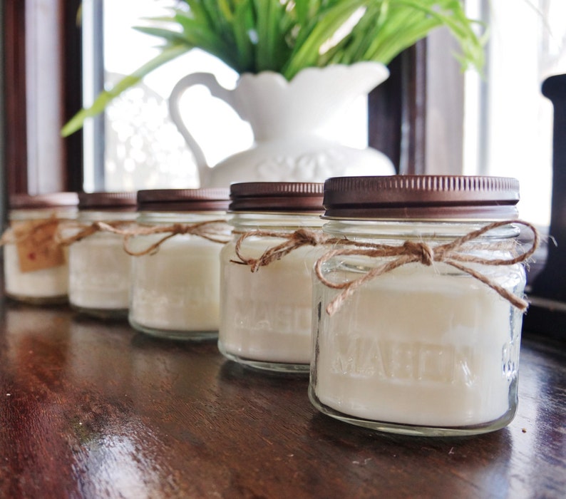 May include: Five glass jars with copper lids and twine tied around the top. The jars are filled with white wax and are arranged in a row on a wooden surface.