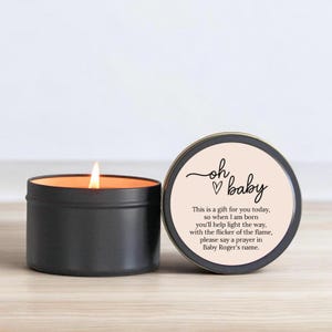 May include: A black candle in a tin with a pink label that says "oh baby" with a heart and the text "This is a gift for you today, so when I am born you'll help light the way, with the flicker of the flame, please say a prayer in Baby Roger's name."