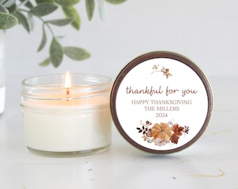 May include: A white candle in a glass jar with a brown and gold label that says "thankful for you Happy Thanksgiving The Millers 2024". The label has a floral design with fall colors.