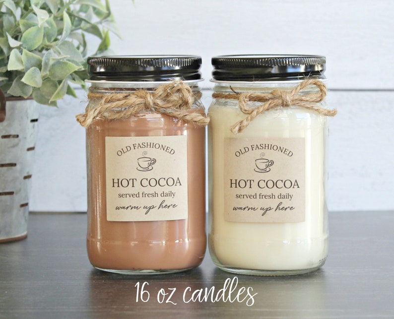 May include: Two 16 ounce glass candle jars with black lids and twine bows. The labels are brown with white text that reads "Old Fashioned Hot Cocoa served fresh daily warm up here".