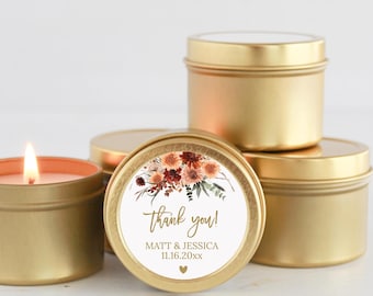 Könnte beinhalten: Eine Gruppe runder Kerzendosen aus goldfarbenem Metall. Eine Dose im Vordergrund hat ein weißes, rundes Etikett mit einer Blumenillustration, dem Text "thank you!", "MATT & JESSICA", "11.16.20xx" und einem kleinen Herz. Eine Kerze brennt.