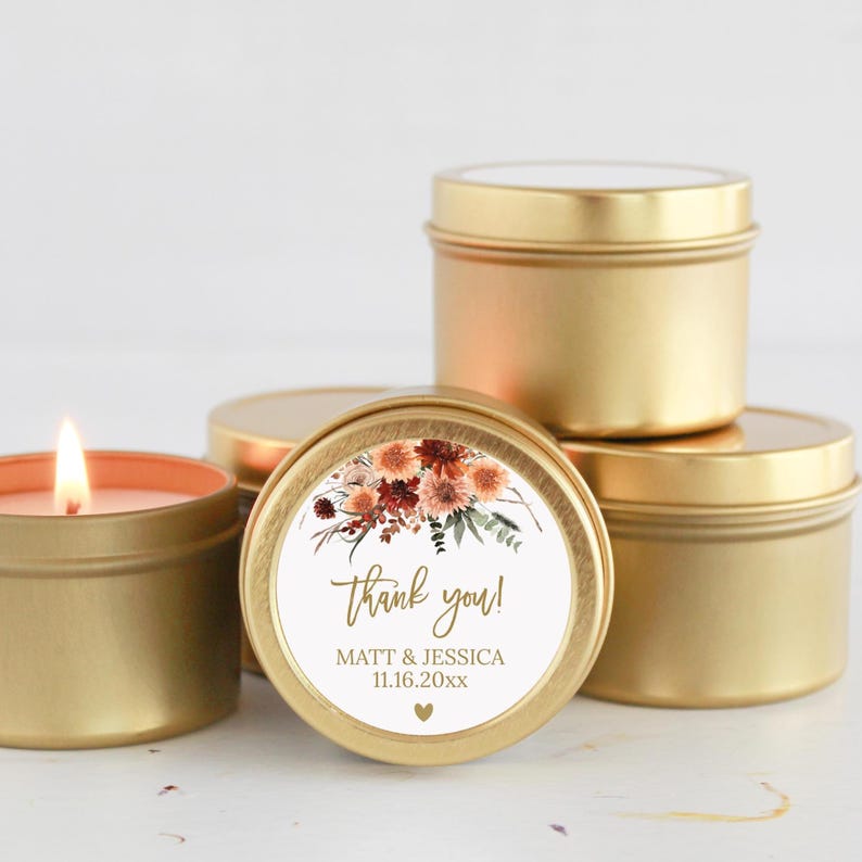 May include: A group of round gold metal candle tins. One tin in the foreground features a white circular label with a floral illustration, the text "thank you!", "MATT & JESSICA", "11.16.20xx", and a small heart. One candle is lit with a visible flame.