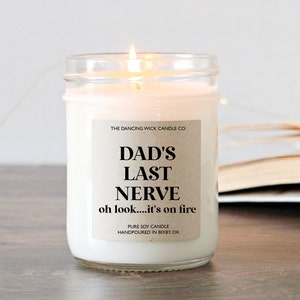 May include: A white soy candle in a glass jar with a brown label that reads "THE DANCING WICK CANDLE CO. DAD'S LAST NERVE oh look....it's on fire PURE SOY CANDLE HANDPOURED IN BIXBY, OK"