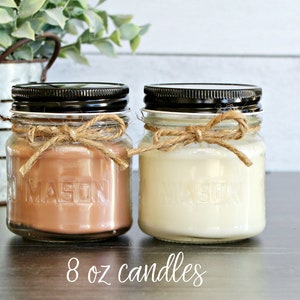 May include: Two 8 ounce candles in glass jars with black lids. The jars have a rustic look with a brown twine bow around the top. One candle is a light brown color and the other is a white color.