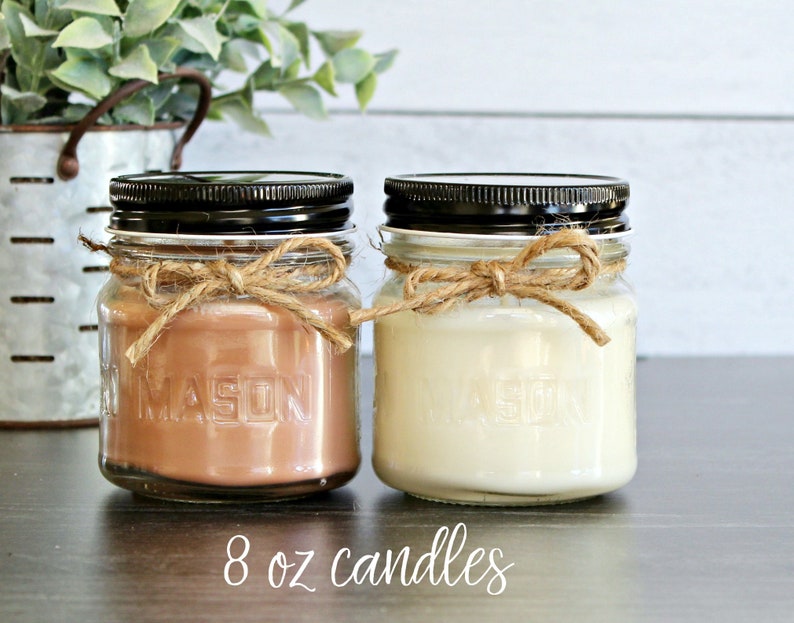 May include: Two 8 ounce candles in glass jars with black lids. The jars have a rustic look with a brown twine bow around the top. One candle is a light brown color and the other is a white color.