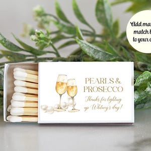 May include: White matchbox with a gold design featuring two champagne flutes and pearls. The text on the box reads "Pearls & Prosecco" and "Thanks for lighting up Whitney's day!"
