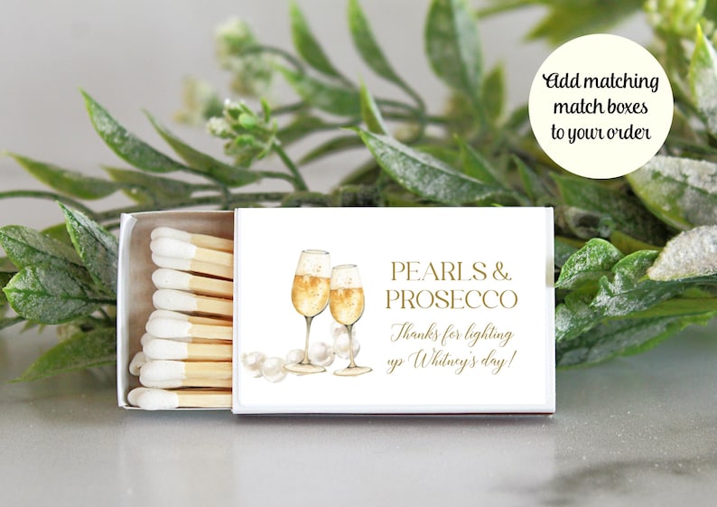 May include: White matchbox with a gold design featuring two champagne flutes and pearls. The text on the box reads "Pearls & Prosecco" and "Thanks for lighting up Whitney's day!"