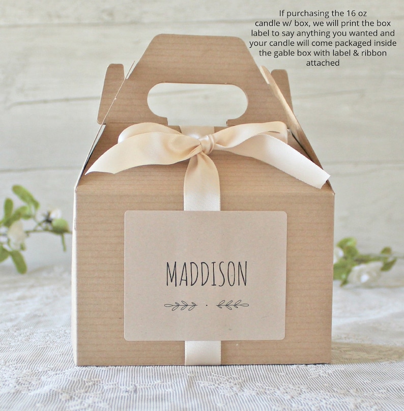 May include: A small, light brown gable box with a handle, tied with a cream-colored ribbon. A rectangular label on the front reads "MADDISON" in black, with leaf accents. The text on the box says, "If purchasing the 16 oz candle w/ box..."