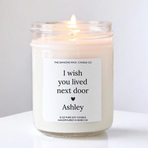 May include: A white soy candle in a clear glass jar with a label that reads "THE DANCING WICK CANDLE CO. I wish you lived next door Ashley 8 OZ PURE SOY CANDLE HANDPOURED IN BIXBY, OK"