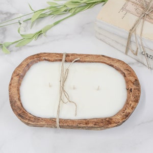May include: An oval-shaped, brown, carved wooden dough bowl candle filled with white wax and featuring three wicks. A piece of jute twine is tied around the center of the bowl. The candle is positioned near a stack of books labeled the dancing wick.