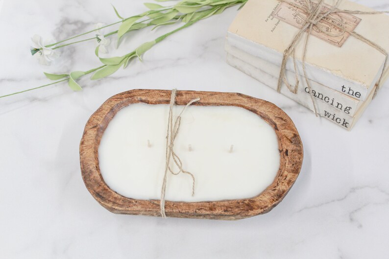 May include: An oval-shaped, brown, carved wooden dough bowl candle filled with white wax and featuring three wicks. A piece of jute twine is tied around the center of the bowl. The candle is positioned near a stack of books labeled the dancing wick.