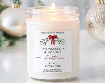 Könnte beinhalten: Eine Kerze im klaren Glas mit einem weißen Etikett, das eine rote Schleife, grüne Zweige und den Text zeigt: THE DANCING WICK CANDLE CO. HAVE YOURSELF A MERRY LITTLE CHRISTMAS LOVE, MACKENZIE 227g PURE SOY CANDLE HANDPOURED IN BIXBY, OK. Die Kerze brennt.