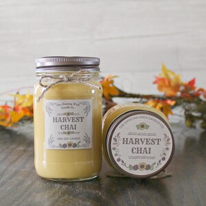 K&ouml;nnte beinhalten: Zwei Glasbeh&auml;lter mit braunen Deckeln und Etiketten. Die Etiketten sind cremefarben mit braunem Text und Bildern von Sonnenblumen. Der Text auf den Etiketten lautet "The Dancing Wick Candle Co." und "Harvest Chai 100% Soy Candle".