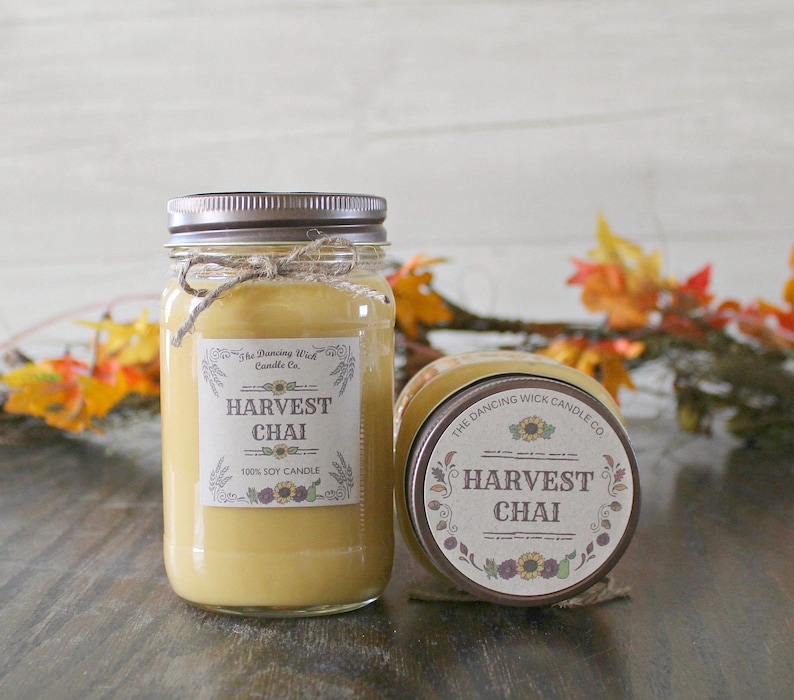 K&ouml;nnte beinhalten: Zwei Glasbeh&auml;lter mit braunen Deckeln und Etiketten. Die Etiketten sind cremefarben mit braunem Text und Bildern von Sonnenblumen. Der Text auf den Etiketten lautet "The Dancing Wick Candle Co." und "Harvest Chai 100% Soy Candle".