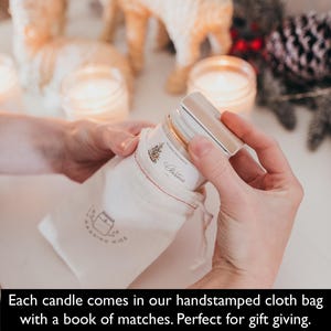 May include: A clear glass jar candle with a wooden lid is being placed into a small white cloth bag. The bag has a red stitched seam and a printed logo. The candle label says "Christmas".