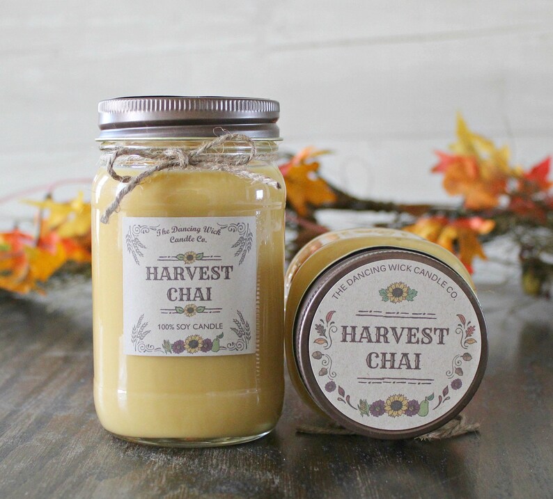 K&ouml;nnte beinhalten: Zwei Glasbeh&auml;lter mit braunen Deckeln und Etiketten. Die Etiketten zeigen "The Dancing Wick Candle Co." und "Harvest Chai 100% Soy Candle".