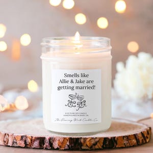 May include: A clear glass jar candle with a white label that reads "Smells like Allie & Jake are getting married!"  The label also includes a small illustration of two champagne glasses with a heart above them. The candle is sitting on a wooden surface.