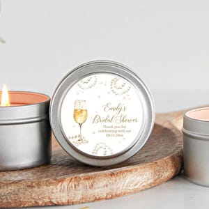May include: Three silver candle tins with white labels. The center label has gold text that reads "Emily's Bridal Shower" and "Thank you for celebrating with us! 08.13.2023". The label also features a watercolor illustration of a champagne flute with a pearl necklace.