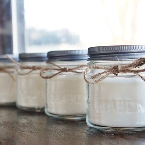 May include: Four glass jars with silver lids and white candles inside. Each jar is tied with a brown twine bow.