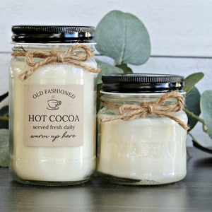 May include: Two glass jars with black lids and twine bows filled with white candle wax. The larger jar has a brown paper label that reads "Old Fashioned Hot Cocoa served fresh daily warm up here".