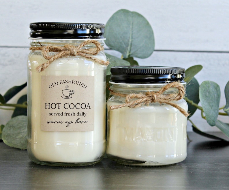May include: Two glass jars with black lids and twine bows filled with white candle wax. The larger jar has a brown paper label that reads "Old Fashioned Hot Cocoa served fresh daily warm up here".