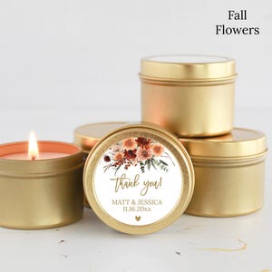 May include: A group of round gold metal candle tins, one of which is lit. A circular label on the front features a floral illustration in shades of orange, red, and brown, with the text "thank you!", "MATT & JESSICA", "11.16.20xx", and a heart.