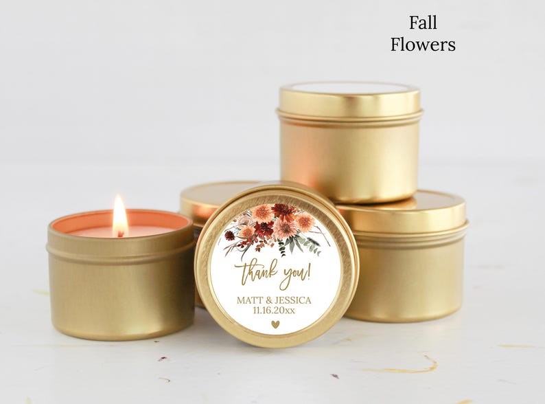 May include: A group of round gold metal candle tins, one of which is lit. A circular label on the front features a floral illustration in shades of orange, red, and brown, with the text "thank you!", "MATT & JESSICA", "11.16.20xx", and a heart.