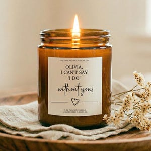 Könnte beinhalten: Eine brennende Kerze in einem bernsteinfarbenen Glas mit einem weißen Etikett, auf dem steht: THE DANCING WICK CANDLE CO. OLIVIA, I CAN'T SAY 'I DO' without you! 9 OZ PURE SOY CANDLE HANDPOURED IN BIXBY, OK. Das Etikett zeigt ein kleines Herz.