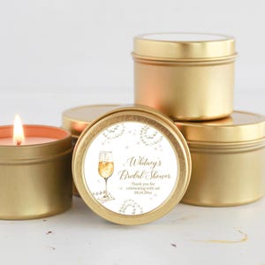 May include: Gold candle tins with a white label that says "Whitney's Bridal Shower" with a champagne glass and pearls. The text on the label also says "Thank you for celebrating with us! 08.14.20xx"