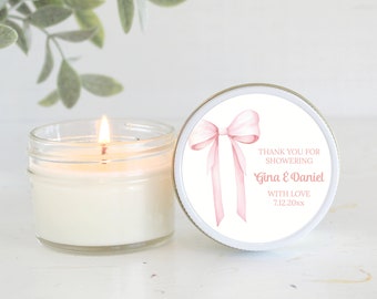 Op de afbeelding: Een witte kaars in een glazen pot met een roze strik op de deksel. De deksel heeft de tekst "Bedankt voor het vieren van Gina & Daniel met liefde 7.12.20xx".