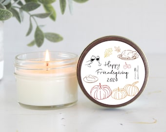 May include: A white candle in a clear glass jar with a brown lid. The lid has a white label with a hand-drawn illustration of a Thanksgiving dinner with the text "Happy Friendsgiving 2024".