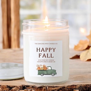 May include: A clear glass jar candle with a white label that reads "THE DANCING WICK CANDLE CO. HAPPY FALL 8 OZ PURE SOY CANDLE HANDPOURED IN BIXBY, OK". The label also features a cartoon illustration of a green pickup truck carrying pumpkins.