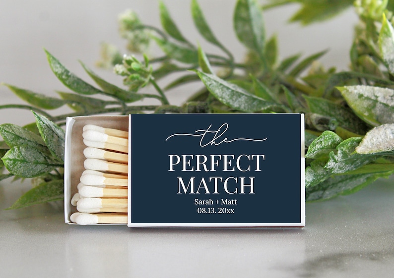 Peut inclure: Une bo&icirc;te d'allumettes rectangulaire avec une couverture bleu marine fonc&eacute; portant le texte blanc "the PERFECT MATCH", les noms "Sarah + Matt" et la date "08.13. 20xx". La bo&icirc;te est entrouverte, r&eacute;v&eacute;lant des allumettes en bois &agrave; bouts blancs.