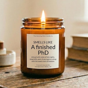 May include: A lit, brown glass jar candle with a white label that reads "SMELLS LIKE A finished PhD." The candle is 9 oz and hand-poured in Bixby, OK. The candle is infused with notes of late nights, long shifts, panic & pure genius.