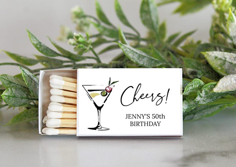 Puede incluir: Caja de cerillas blanca con una ilustraci&oacute;n en blanco y negro de una copa de martini con aceitunas. El texto "Cheers! Jenny's 50th Birthday" est&aacute; impreso en la caja de cerillas.